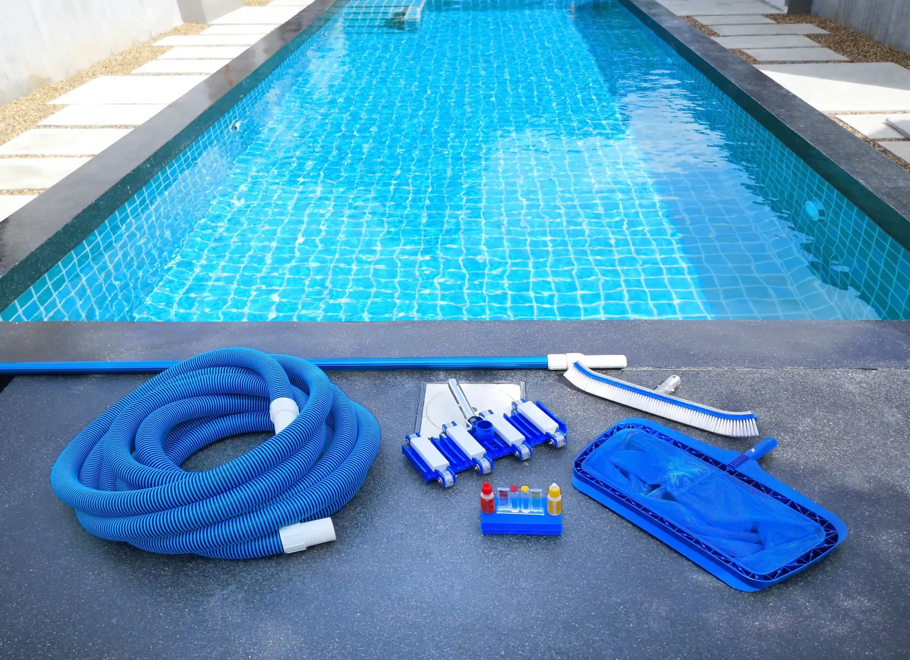 Pool vacuum hose, brush, test kit, and skimmer net laid out beside a clean residential pool